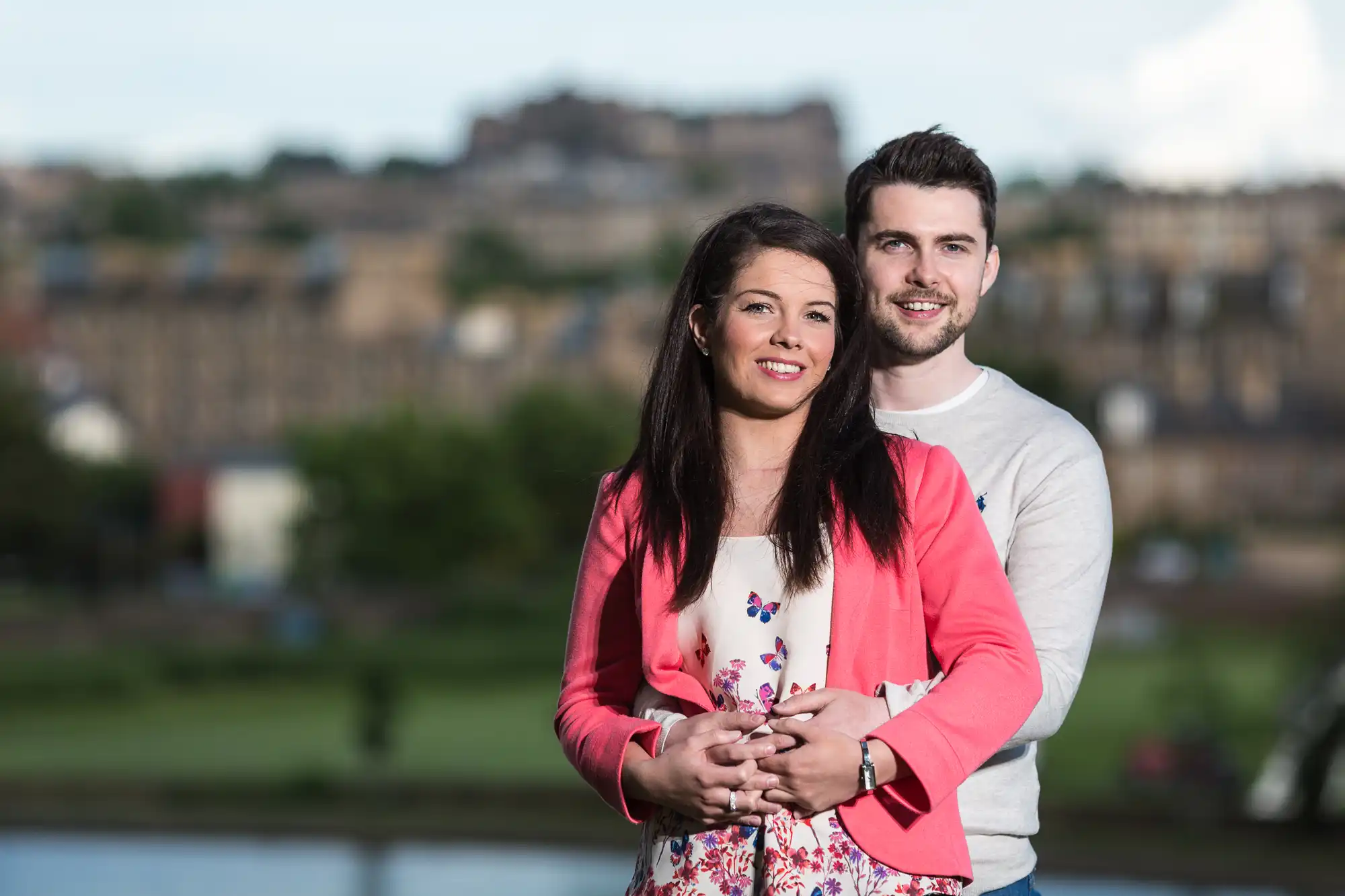A young couple embracing and smiling outdoors with a blurred cityscape and hilltop castle in the background.