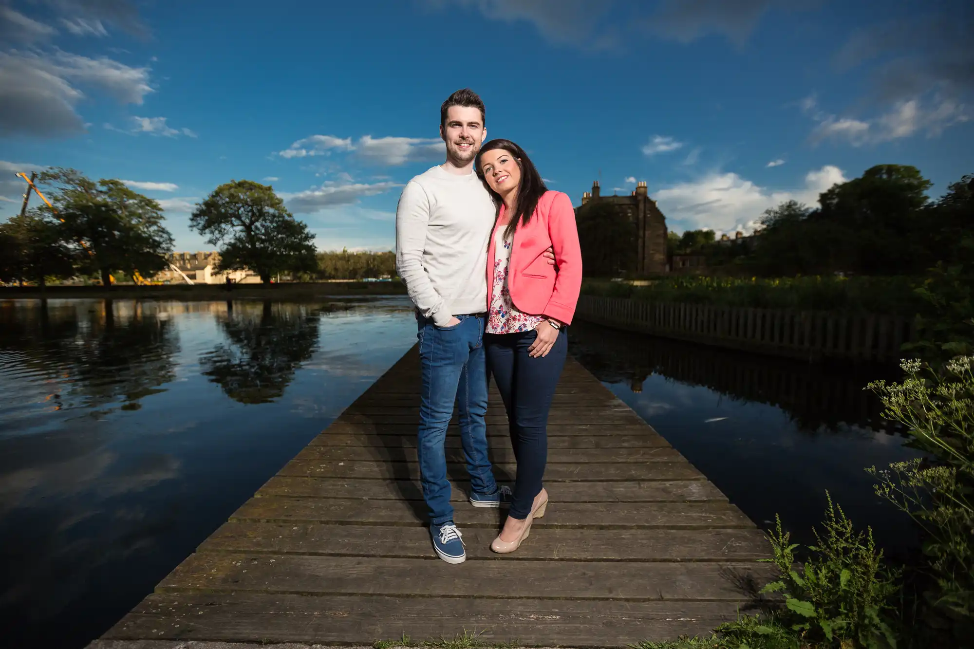 A smiling couple stands together on a wooden pier beside a calm lake, with lush greenery and a cloudy blue sky in the background.