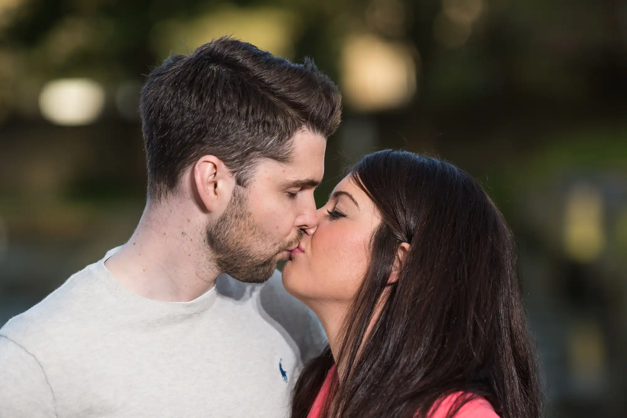 A man and a woman kissing each other on the lips in a park, with trees blurred in the background.