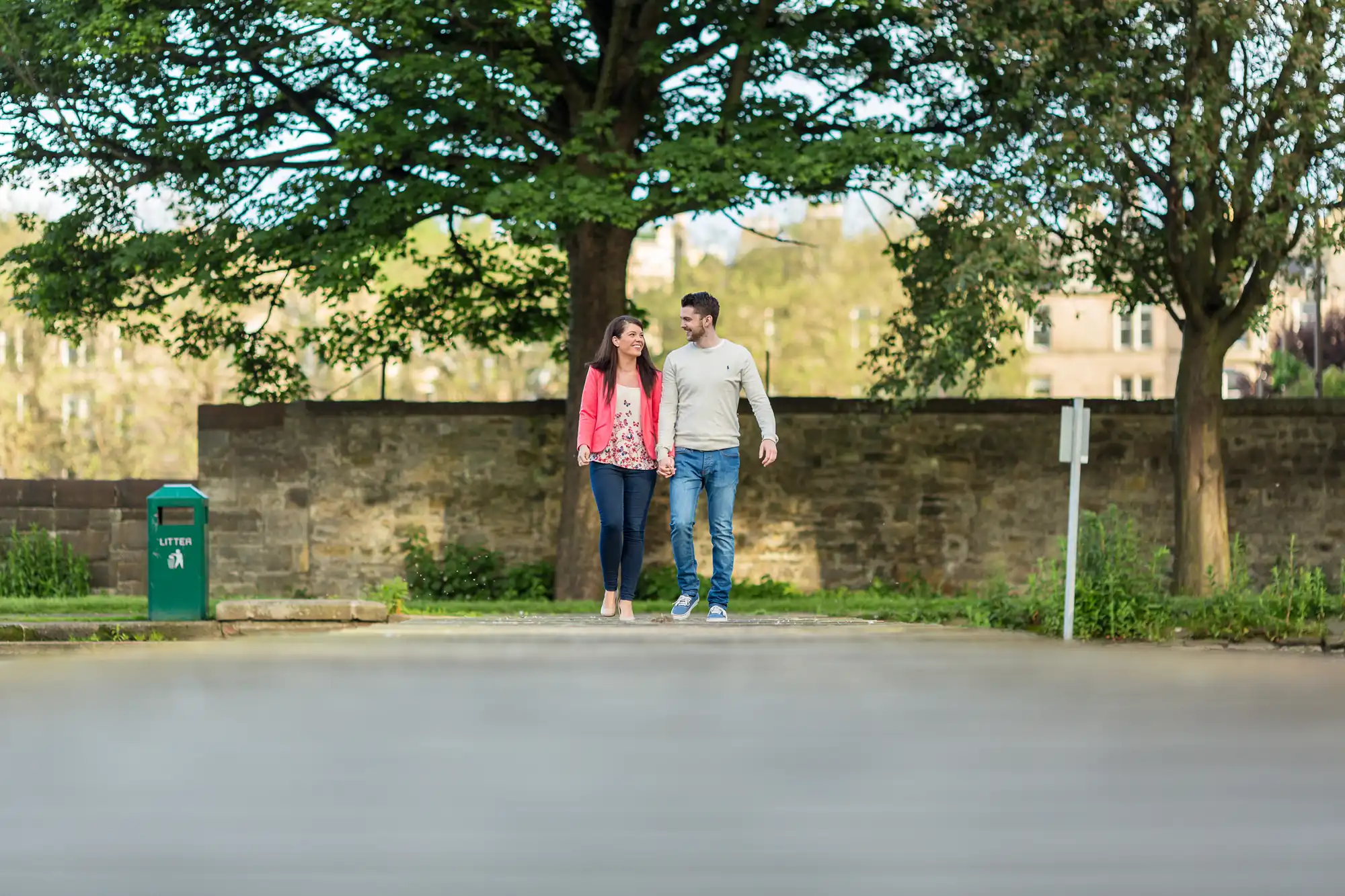 A couple walking hand in hand on a sunny suburban street, smiling and conversing, with trees and a stone wall in the background.