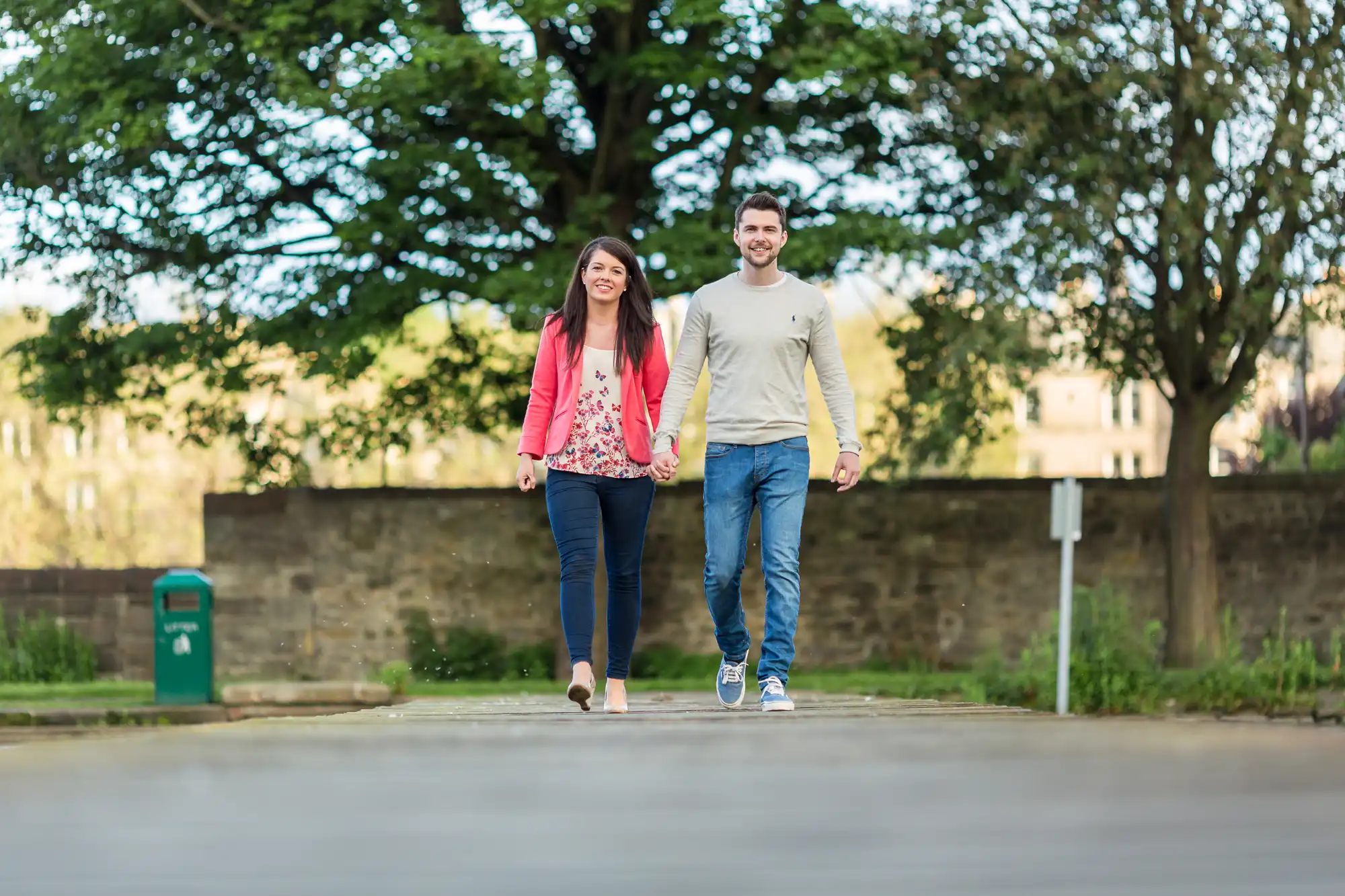 A young couple holding hands and smiling while walking on a paved path in a park with trees in the background.