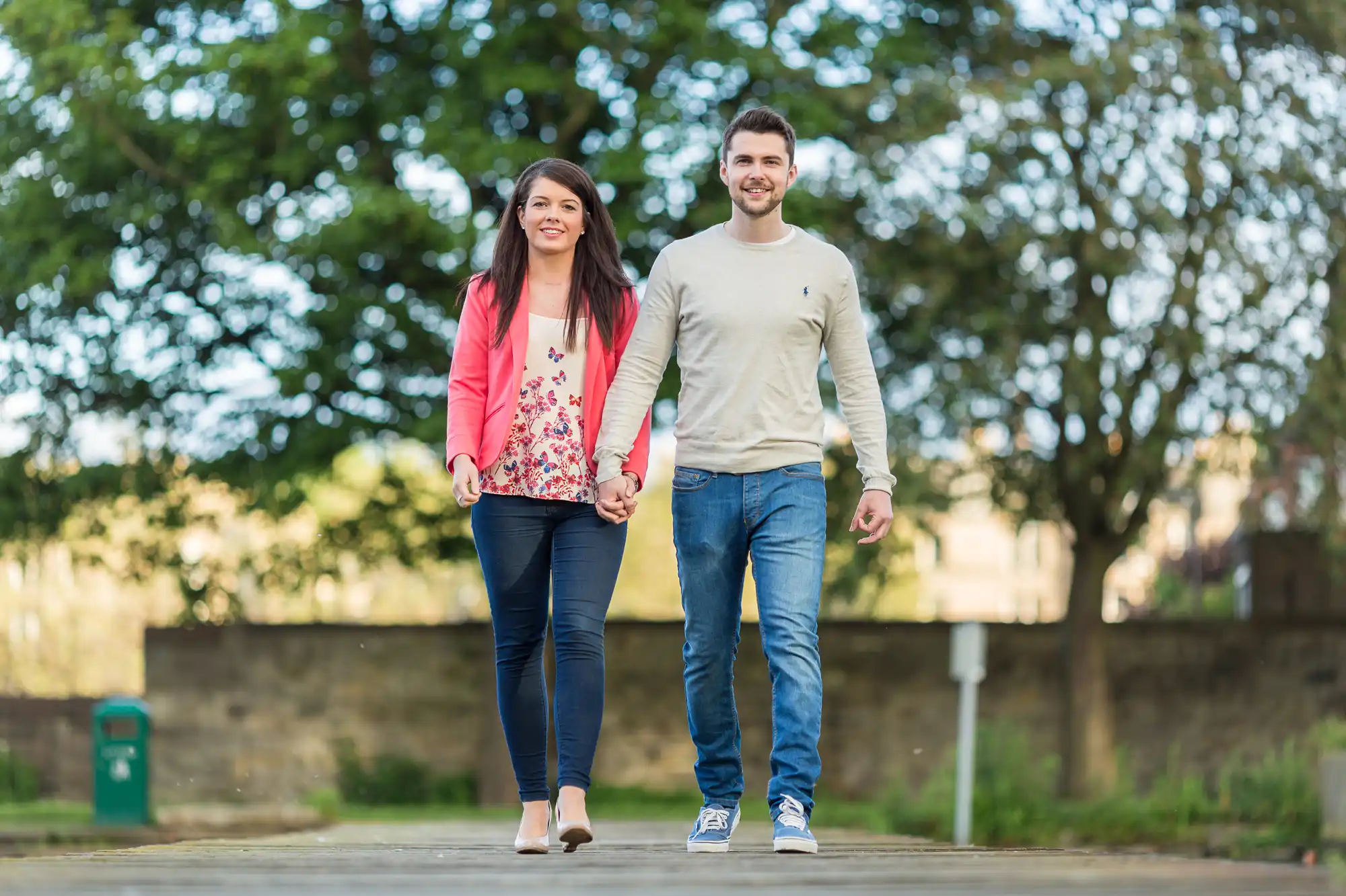 A young couple happily holding hands while walking on a paved path in a park, smiling at the camera.