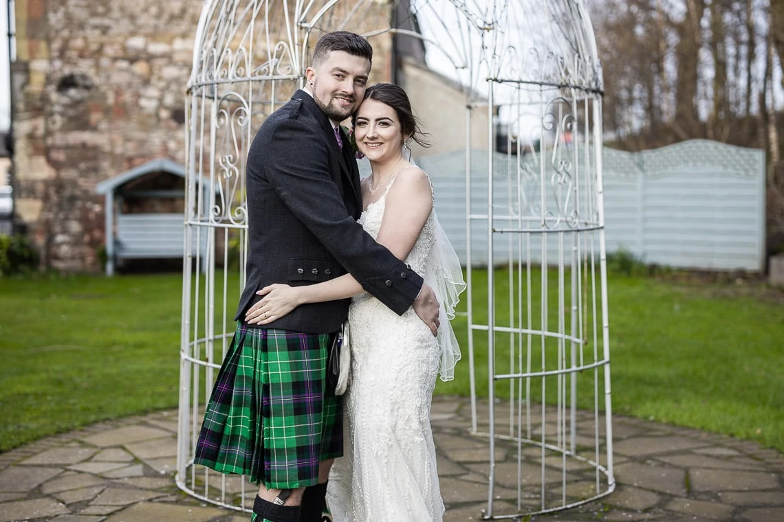 Newlyweds embrace in front of the bird cage in the garden.