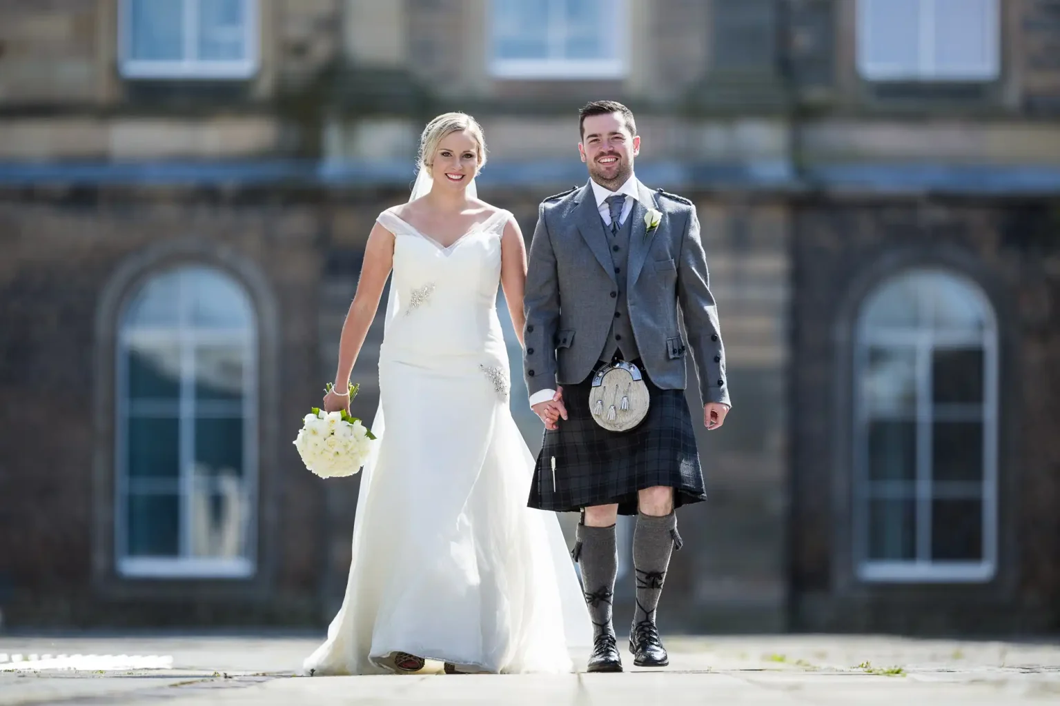 A couple dressed in wedding attire, the woman in a white gown holding a bouquet, and the man in a kilt and jacket, walk hand-in-hand outside a building with large windows.