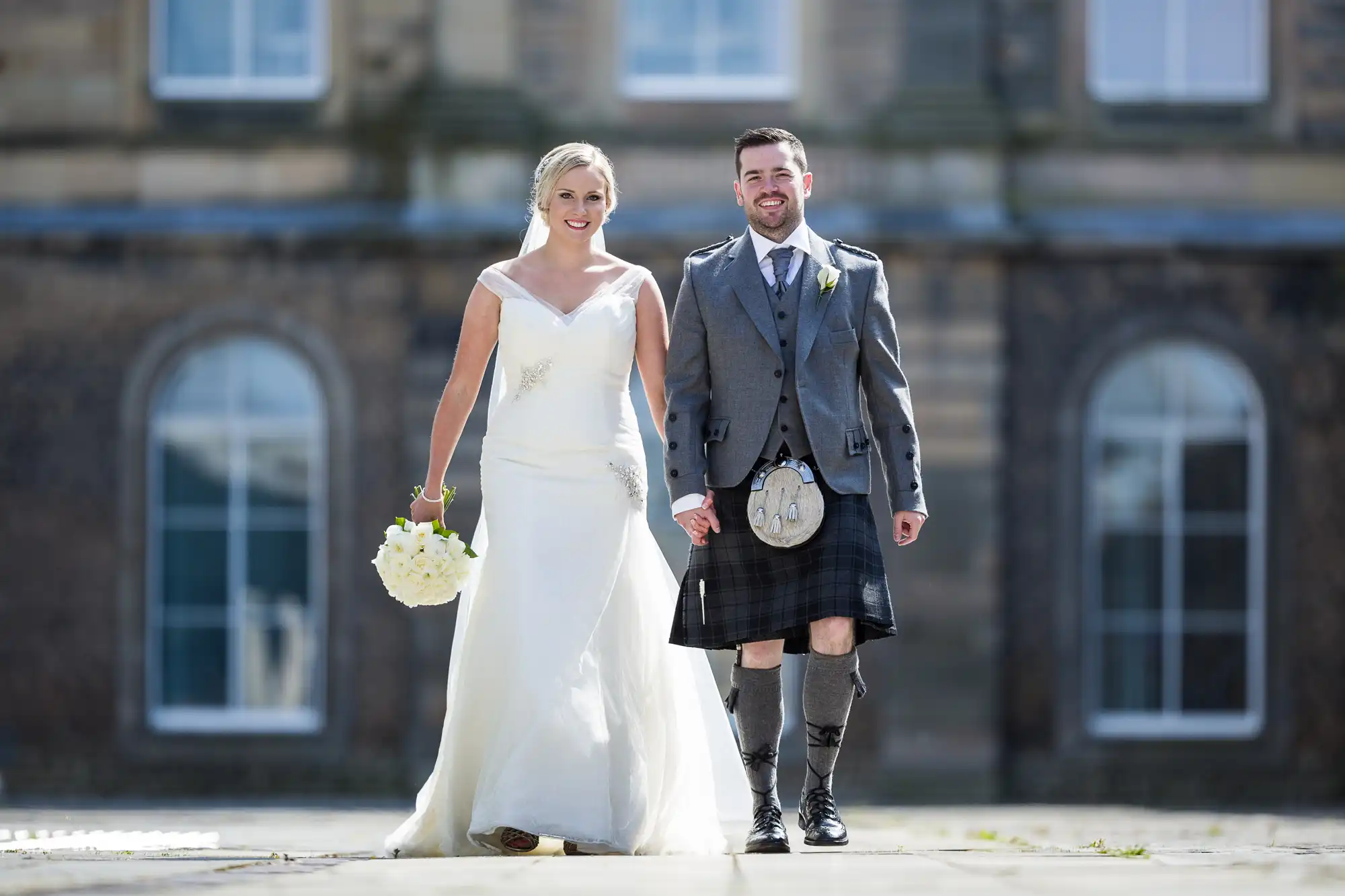 A couple dressed in wedding attire, the woman in a white gown holding a bouquet, and the man in a kilt and jacket, walk hand-in-hand outside a building with large windows.