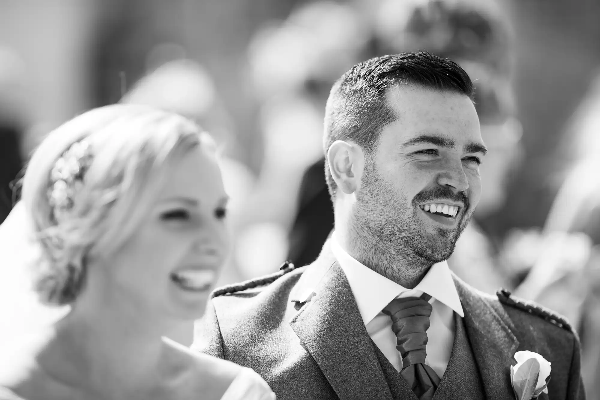 A smiling bride and groom in wedding attire share a joyful moment. The groom wears a suit with a tie, and the bride has blonde hair and a veil. The image is in black and white.