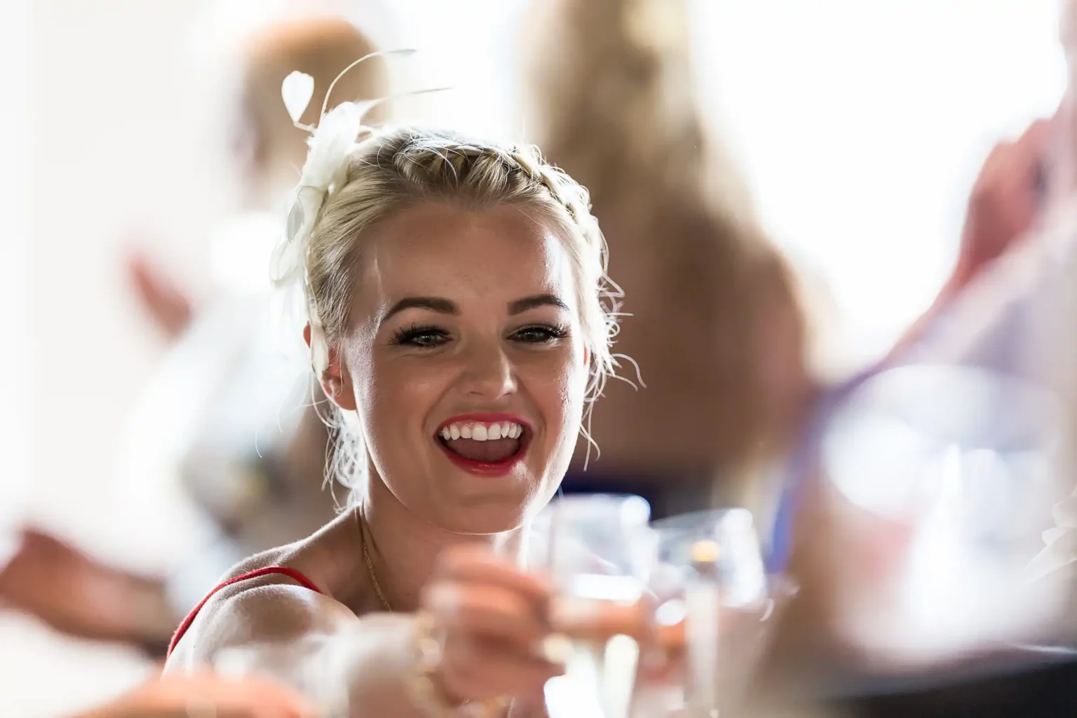 A woman, dressed in formal attire and a white headpiece, smiling and reaching out with a glass in hand at a social gathering.