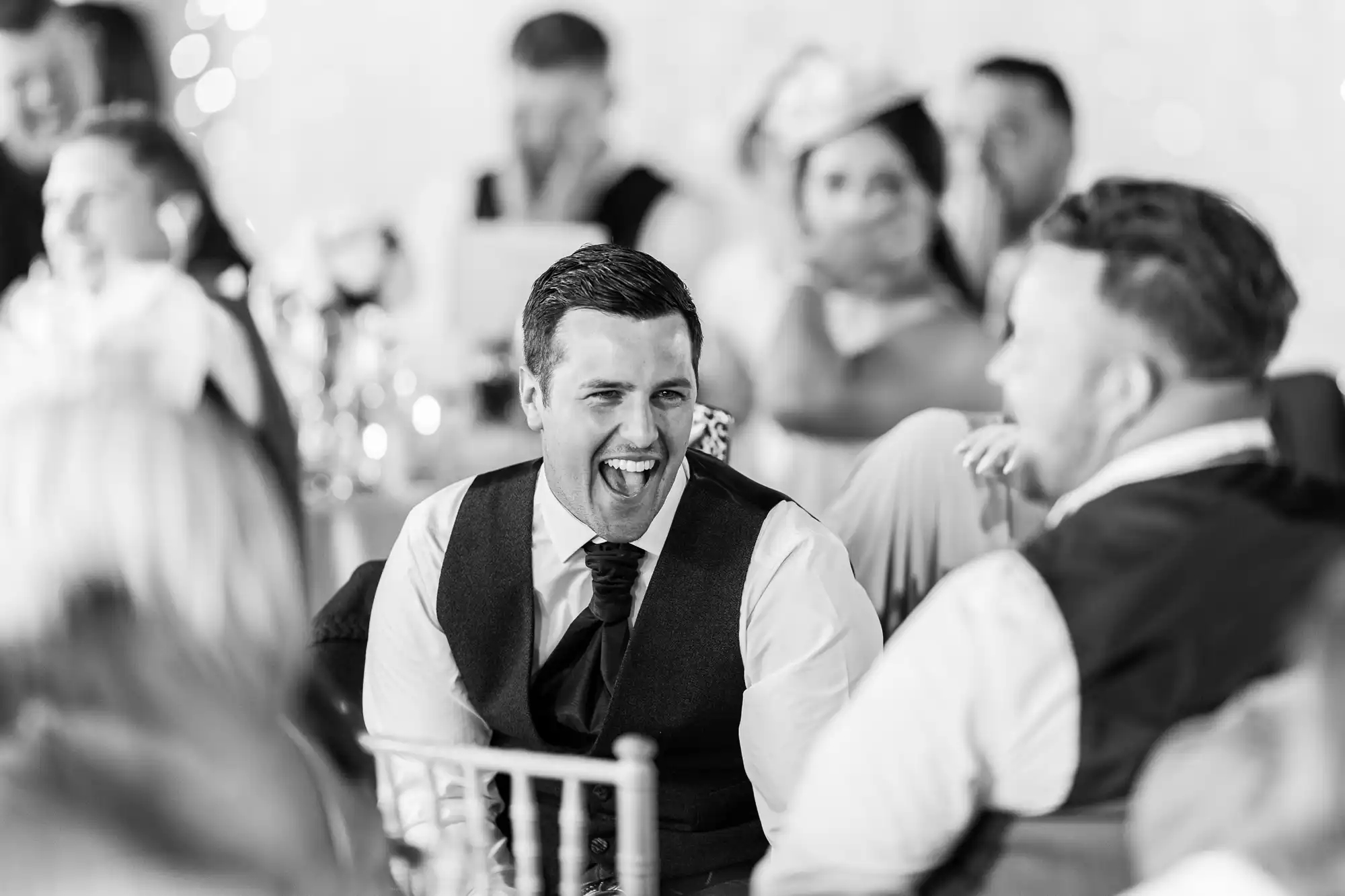 A man in formal attire is laughing heartily while seated, surrounded by other people who are also dressed formally.