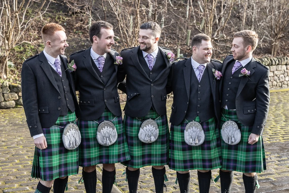 Five men in traditional scottish kilts and jackets, laughing and embracing each other at an outdoor gathering.