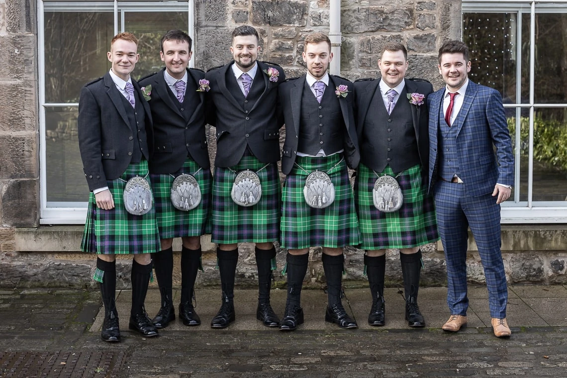 Six men in traditional scottish kilts and one man in a suit posing together in front of a stone building.