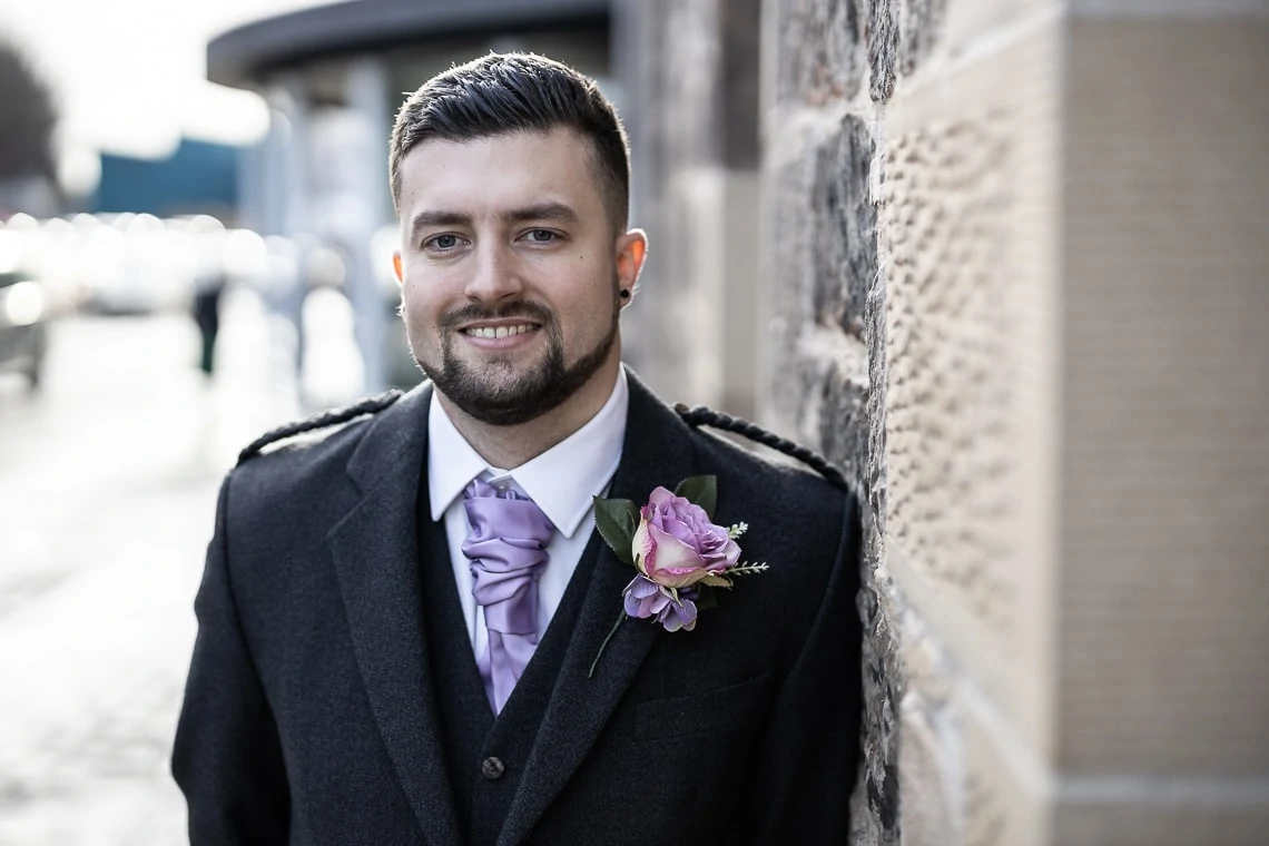 A man in a black suit and lavender tie with a matching boutonniere smiles outdoors near a brick wall.