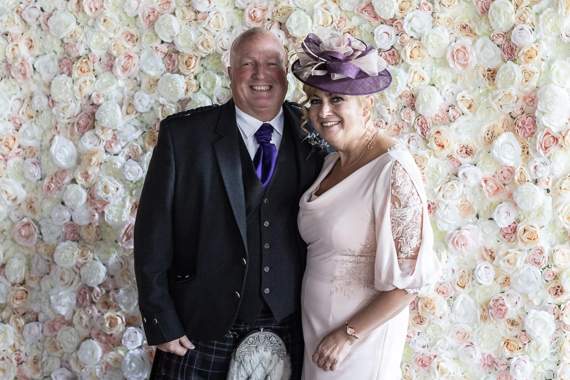 A smiling couple dressed in formal wedding attire, with the man in a kilt and the woman in a pastel dress, standing in front of a floral wall.