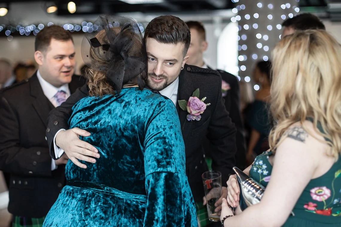 A bride in a teal dress and black fascinator hugs a groom in a gray suit, while a woman with a floral dress holds a notebook nearby, in a room with string lights.
