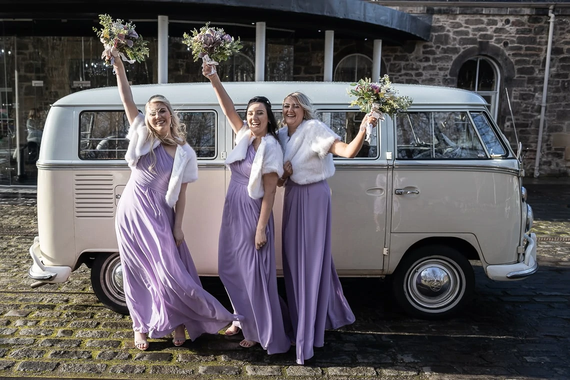 Three bridesmaids in purple dresses and white fur stoles cheerfully pose with bouquets beside a vintage white van.