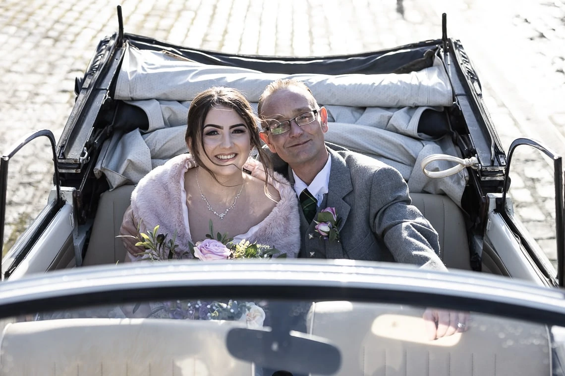A bride and her father smiling in the backseat of a convertible car, dressed elegantly for a wedding.