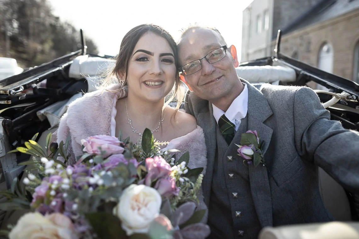 A bride in a pink shawl and a groom in a gray suit smiling inside a vintage convertible car, holding a bouquet of flowers.
