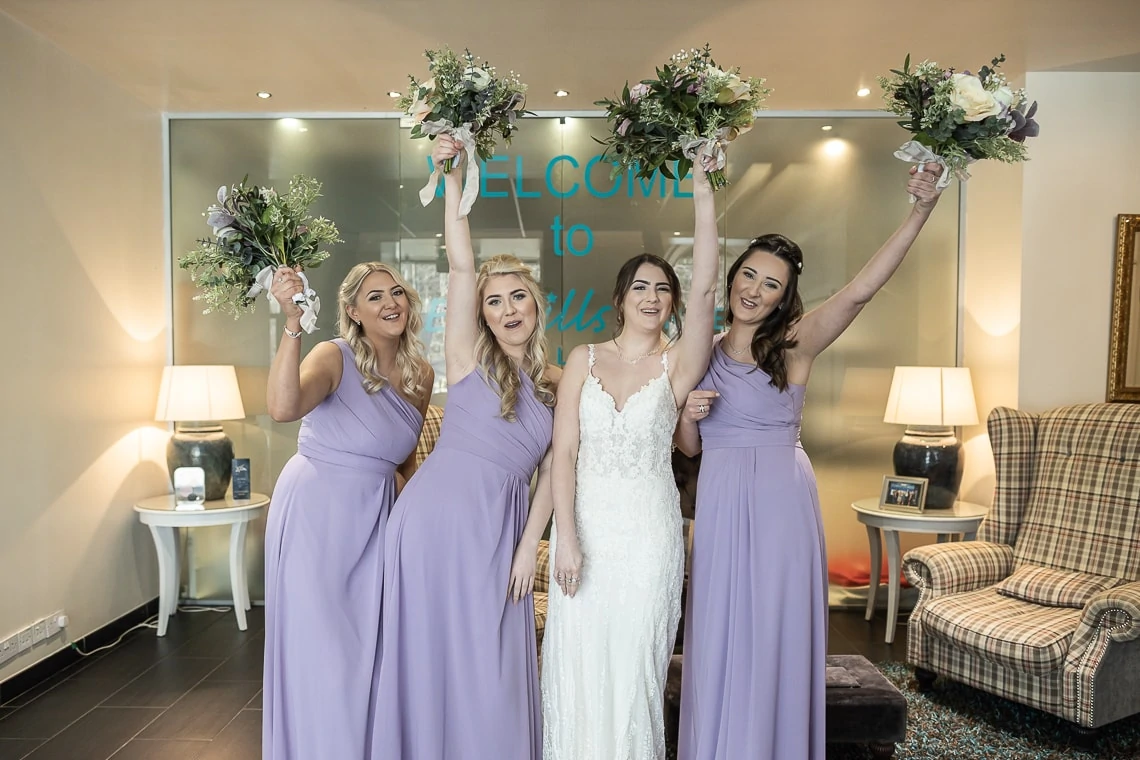 Four women in lavender dresses raising bouquets, celebrating with the bride in a white dress in a hotel lobby.