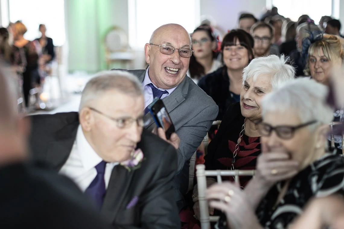 A group of elderly guests smiling and enjoying themselves at a social event, one man laughing while holding a smartphone.