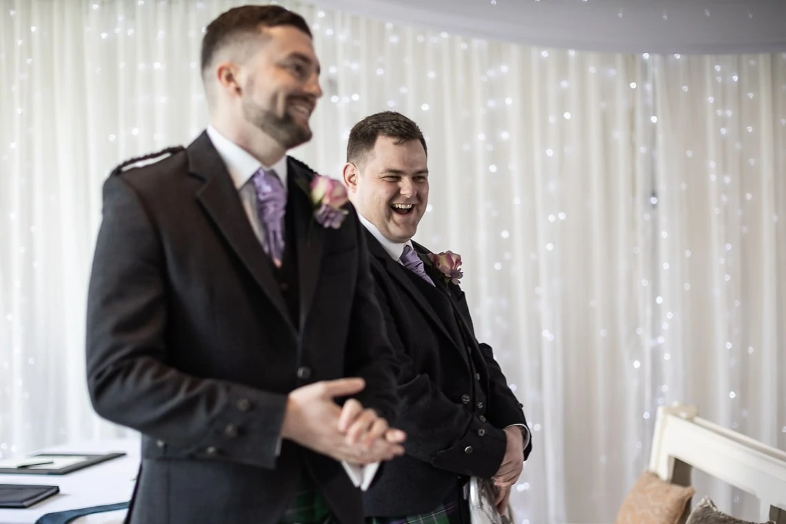 Two men in formal attire, one in a military uniform and the other in a kilt, laugh joyfully at a wedding ceremony.