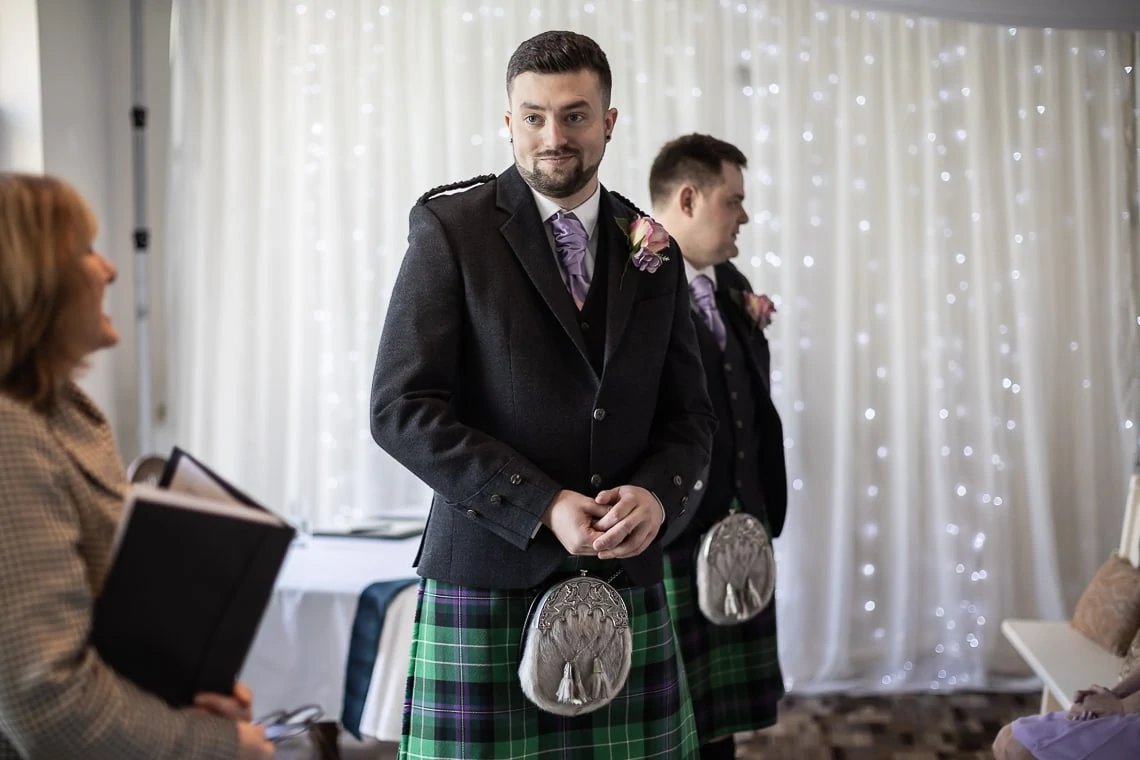 Two men in traditional scottish kilts and jackets at a formal event, standing in front of a curtain with fairy lights. one man looks towards the camera.