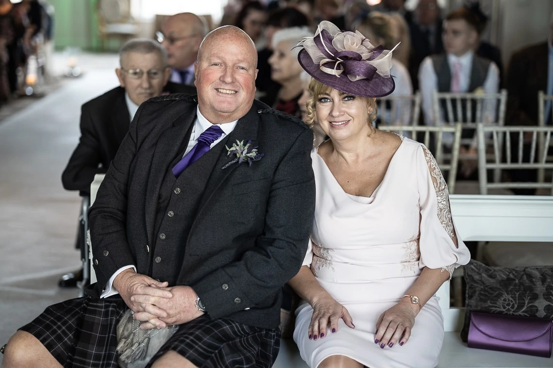 A smiling couple dressed in formal attire at a wedding ceremony, the man in a kilt and the woman wearing a hat with a large purple flower.