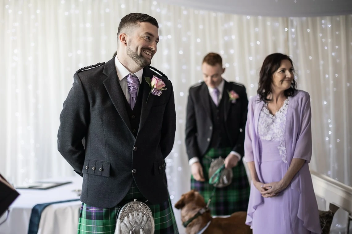 A groom in a tartan kilt and jacket smiles during a wedding ceremony, with a bridesmaid and another man in kilts in the background.