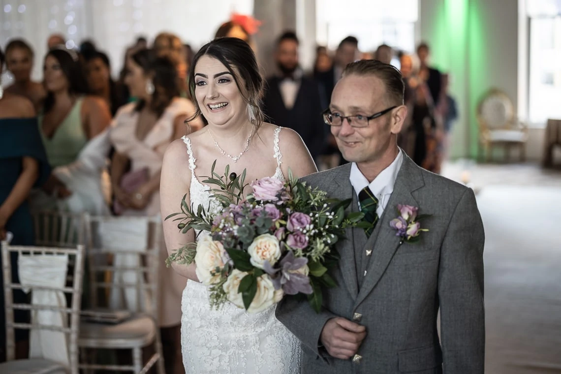 A bride in a white gown, smiling widely, walks down the aisle with her father who is wearing glasses and a suit, both holding a bouquet of flowers.
