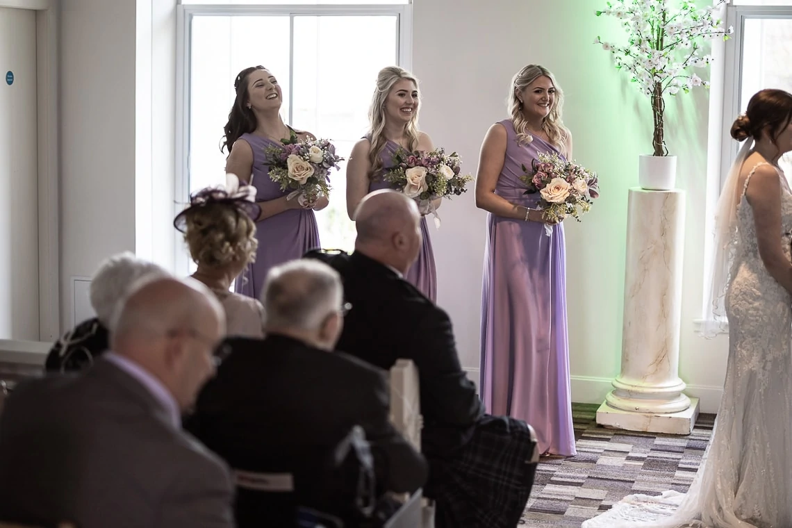 Three bridesmaids in lavender dresses holding bouquets smile attentively at a bride during a wedding ceremony indoors.
