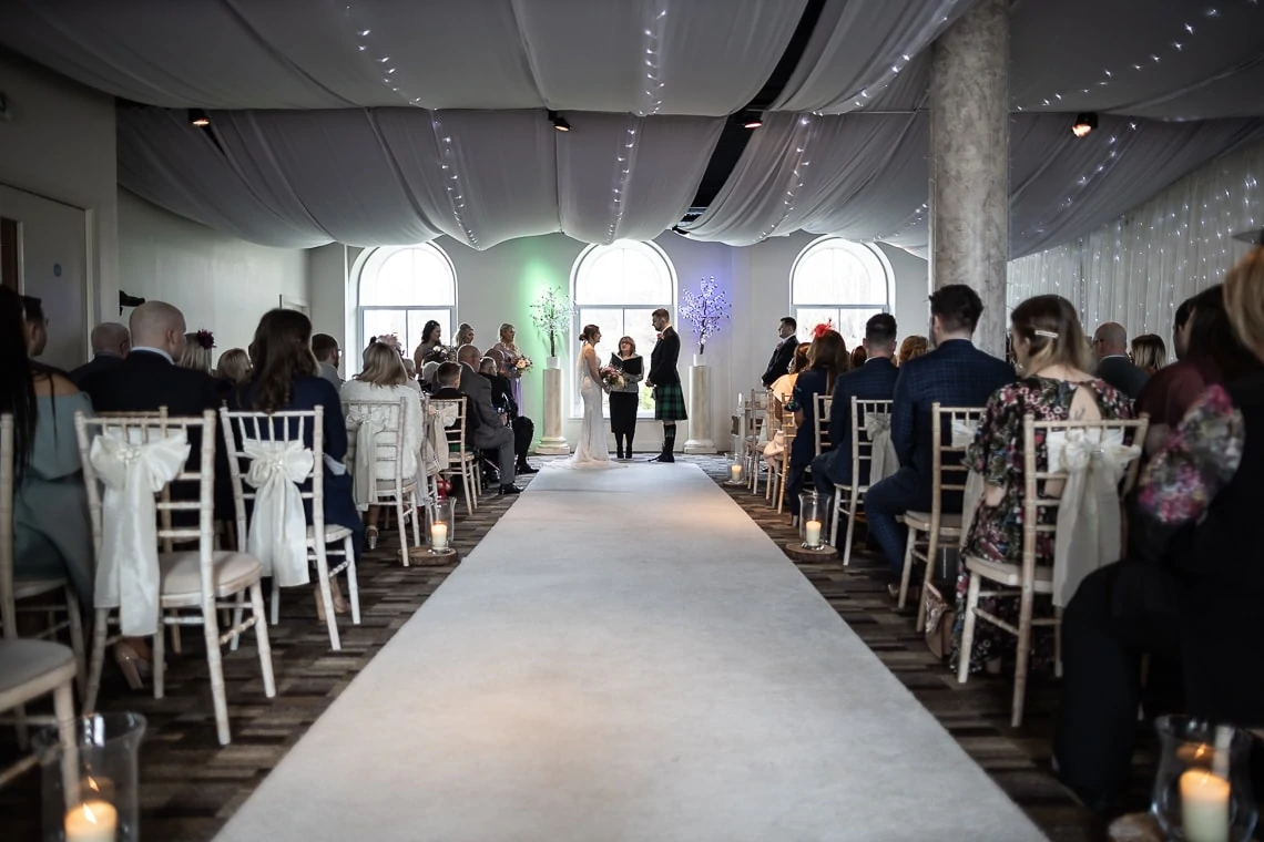 A wedding ceremony in an elegant indoor venue with white chairs, guests seated on either side, and a couple standing at the altar under soft lighting.