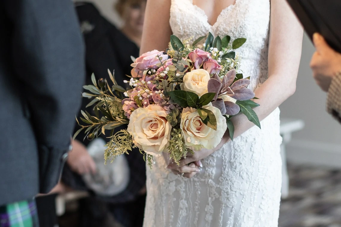Bride in a lace dress holding a bouquet of roses and mixed flowers at a wedding ceremony, with guests partially visible in the background.