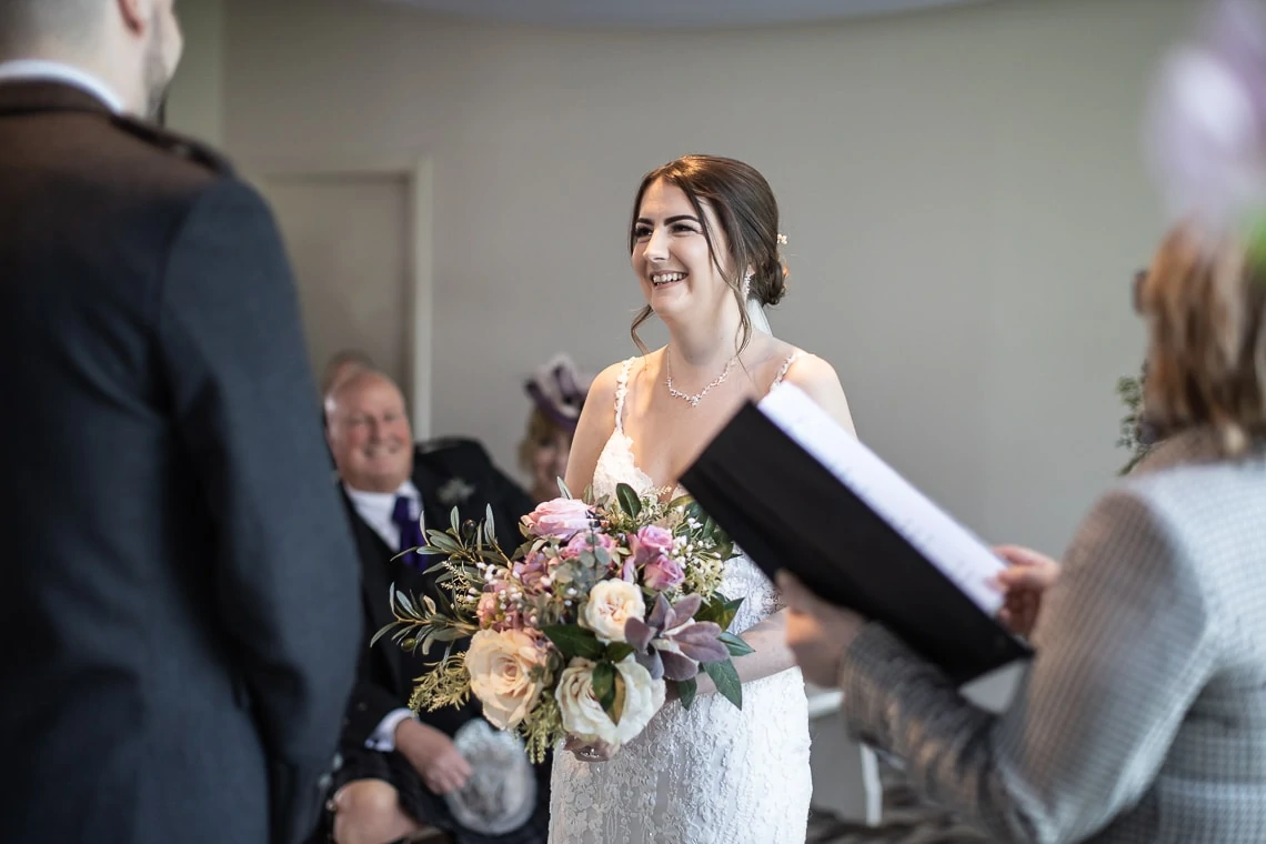 A radiant bride holding a bouquet laughs joyfully during her indoor wedding ceremony, with guests looking on.