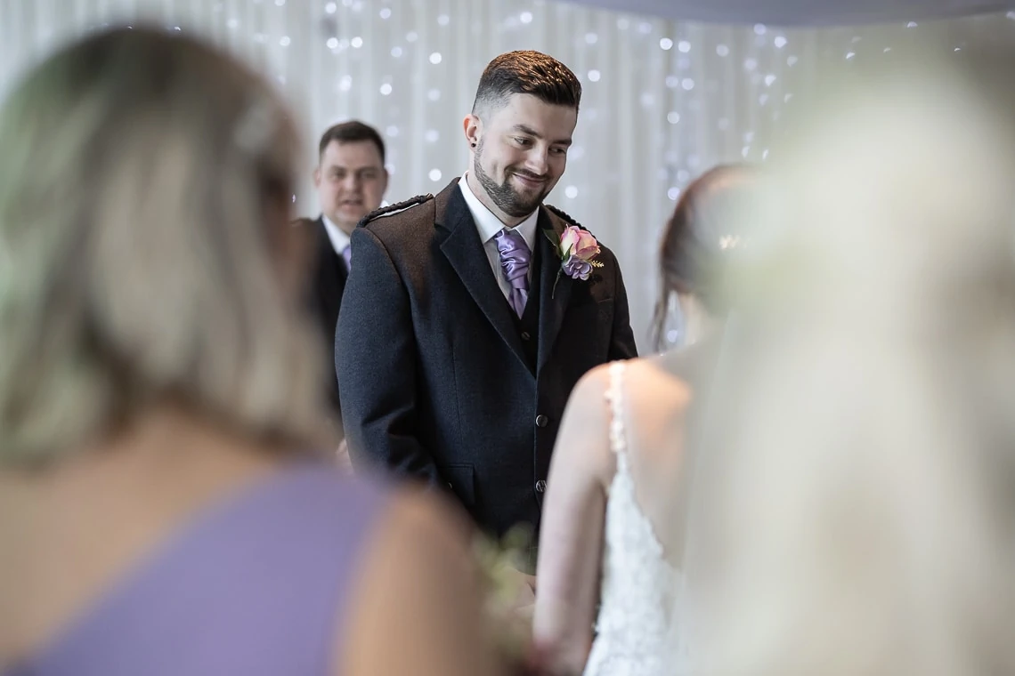 A groom in a gray suit smiles at a bride in a white dress during a wedding ceremony, with guests in the foreground.
