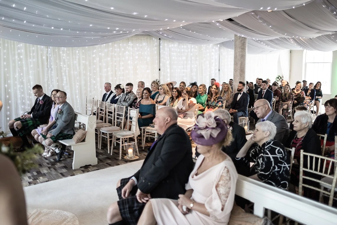Guests seated in rows at a wedding ceremony inside a draped, elegantly lit hall, with some attendees dressed in kilts.