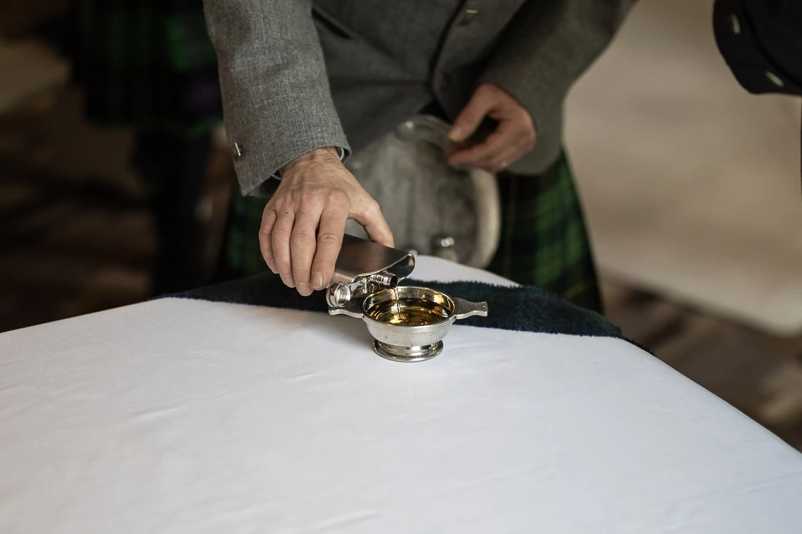 A person in a kilt pours whisky from a flask into a silver quaich placed on a white tablecloth.