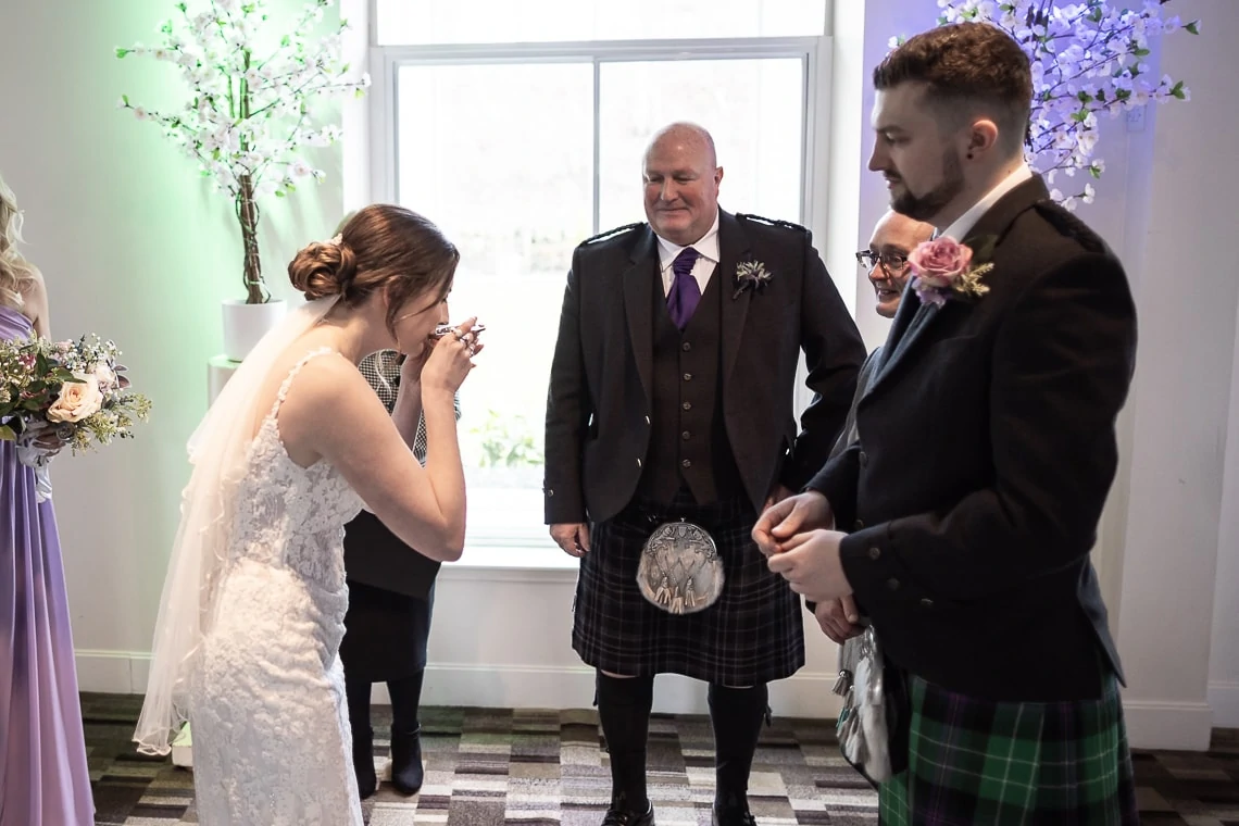 A bride in a white dress and a man in a kilt watch another woman emotional during a wedding ceremony indoors.