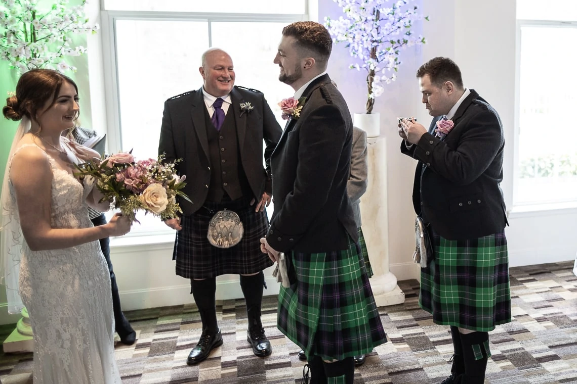 Bride and three men in scottish kilts, one adjusting a lapel flower, smiling and interacting joyfully in a room decorated with floral arrangements.