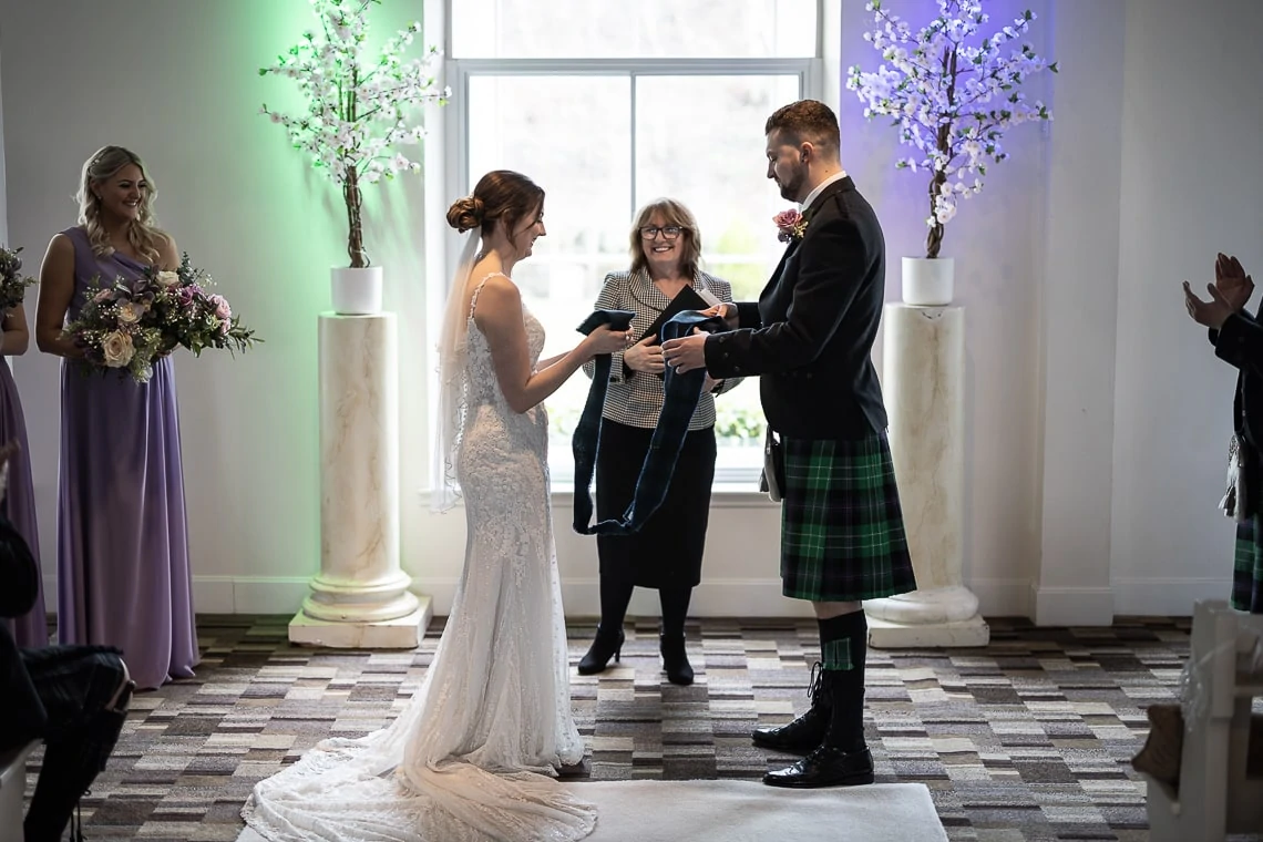 Bride and groom exchanging rings at their wedding ceremony, officiant and guests in attendance, groom wearing a kilt.