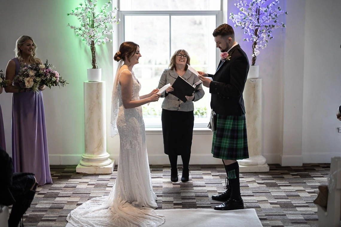 A bride and groom exchanging vows in front of an officiant at a wedding ceremony, with two bridesmaids watching. the groom is wearing a kilt.