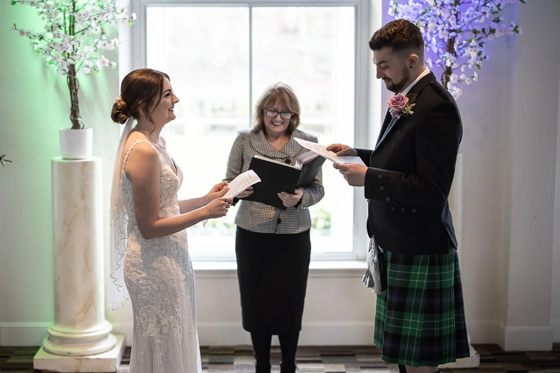 A bride and groom exchanging vows in front of an officiant, with the groom wearing a kilt, in a room with floral decorations.