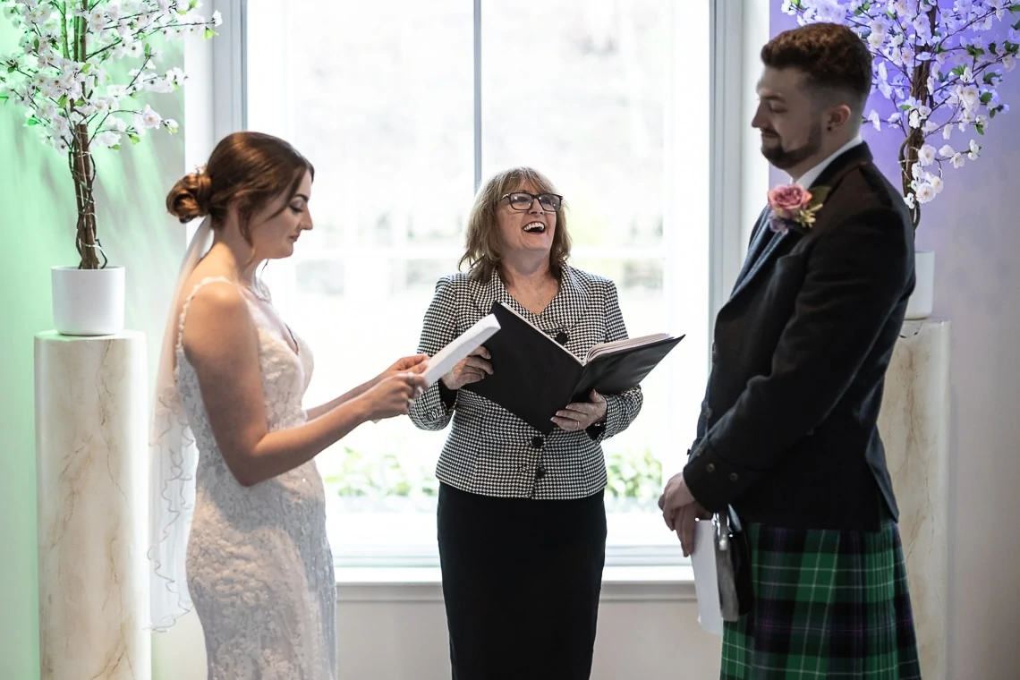 A bride and groom exchanging vows with an officiant holding a book, standing by a window with floral decorations.