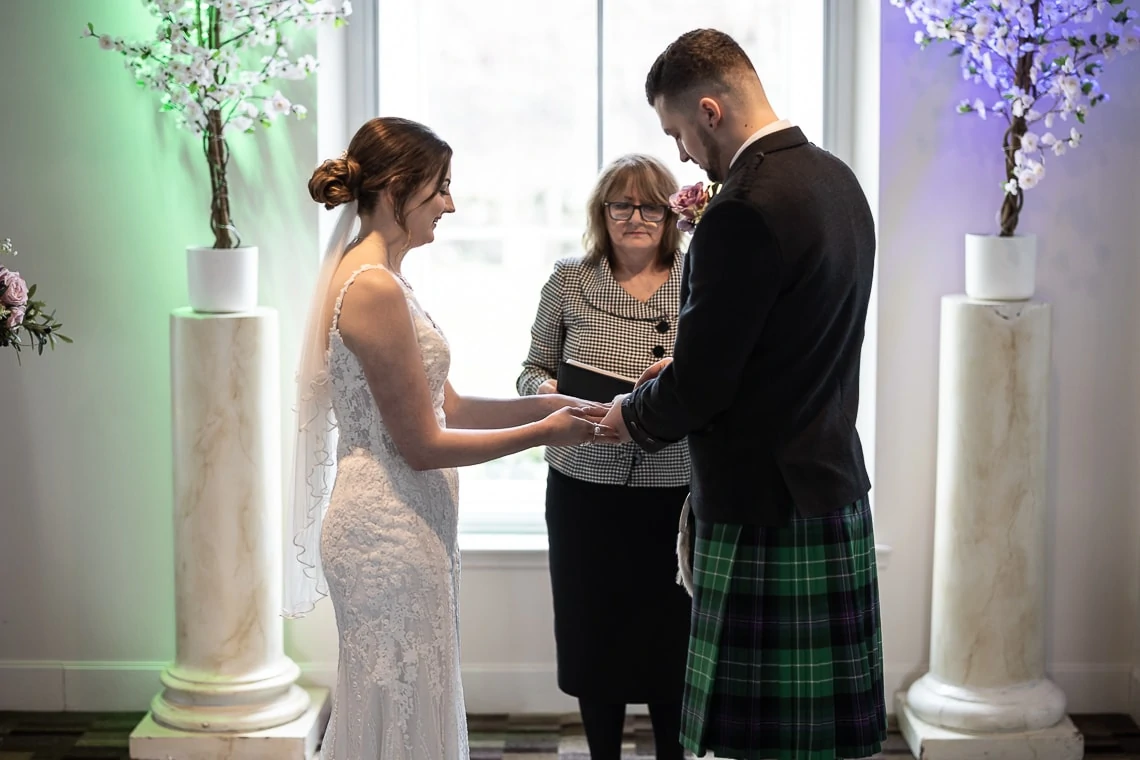 A bride in a white dress and a groom in a kilt exchanging vows with an officiant in between, inside a room decorated with floral arrangements.