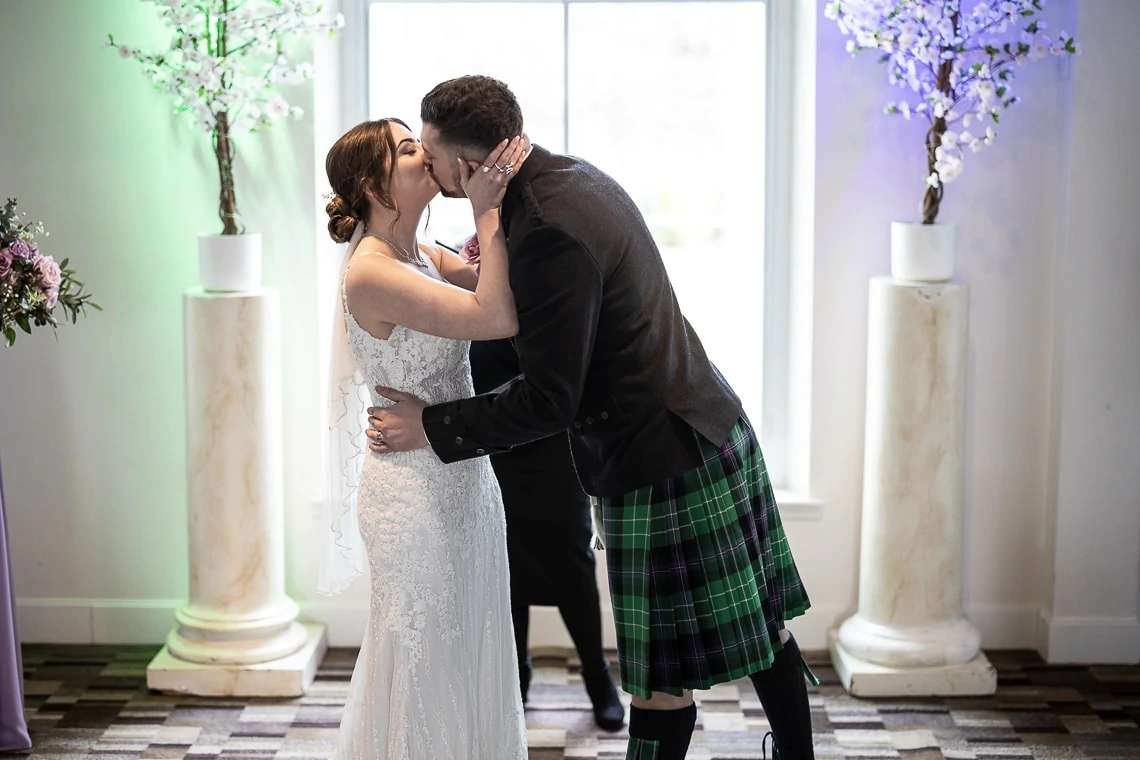 A bride and groom kissing passionately, the groom wears a kilt and the bride a white dress, in a room with floral decorations and daylight streaming through a window.