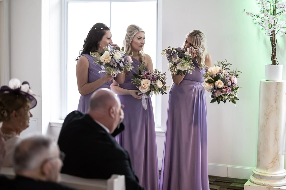 Three bridesmaids in purple dresses holding bouquets at a wedding ceremony, whispering to each other, with guests seated nearby.