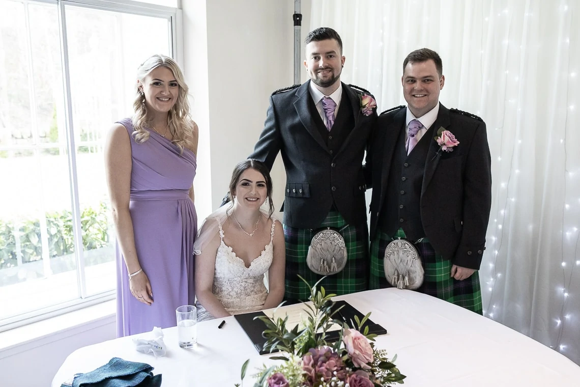 A bride seated at a table with three guests standing beside her: two men in kilts and a woman in a purple dress, in a room with a bright window background.