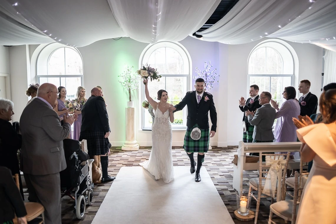 A newlywed couple walks down the aisle, the bride in a white dress and the groom in a kilt, with guests clapping around them in a brightly lit room.