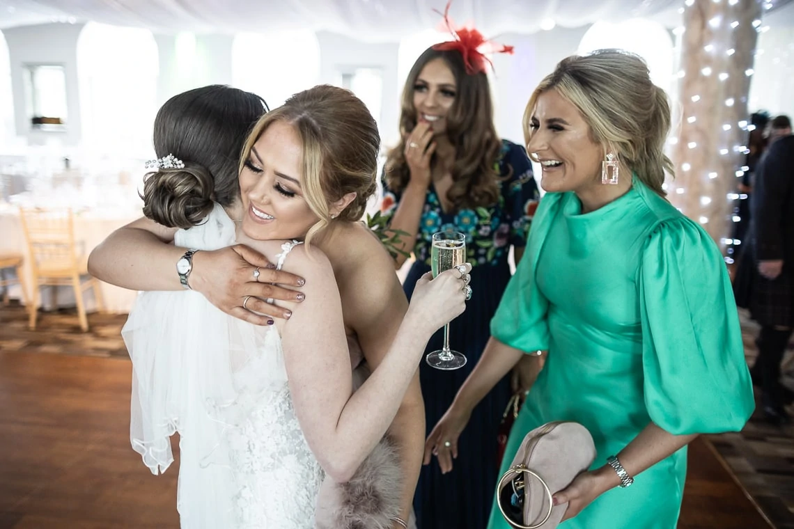A bride in a white dress hugging a young girl, while two women, one holding a champagne glass, joyfully watch.