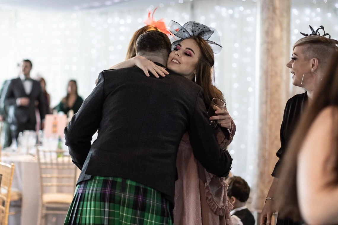 Woman in a fascinator embracing a man in a tartan kilt at a festive event, with guests in the background.