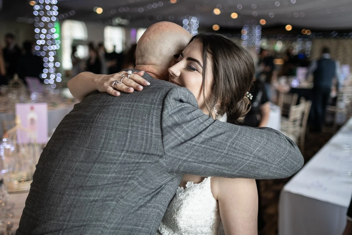 A bride hugs a bald man at a wedding reception, her expression joyful; they are surrounded by guests and tables decorated for the event.