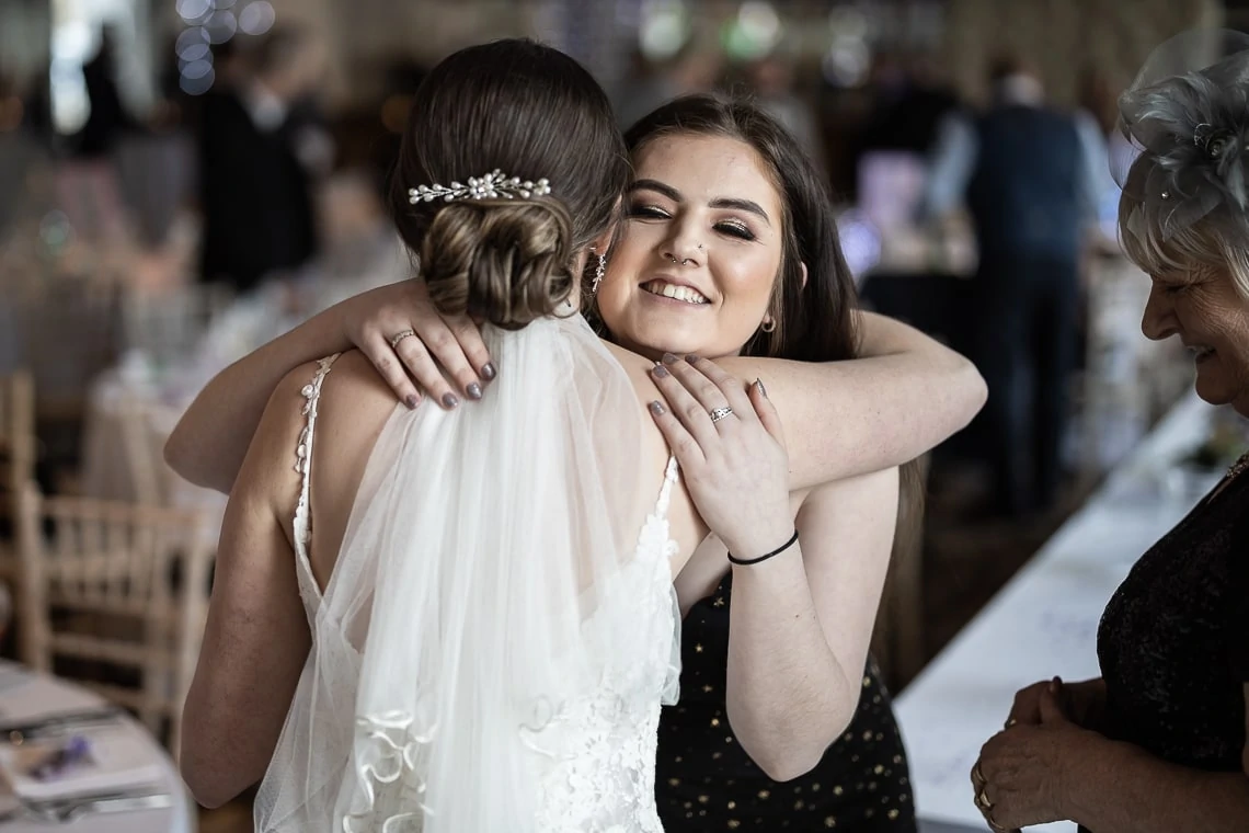 A joyful bride in a white dress, hugging a smiling woman in a black and gold dress at a wedding reception.