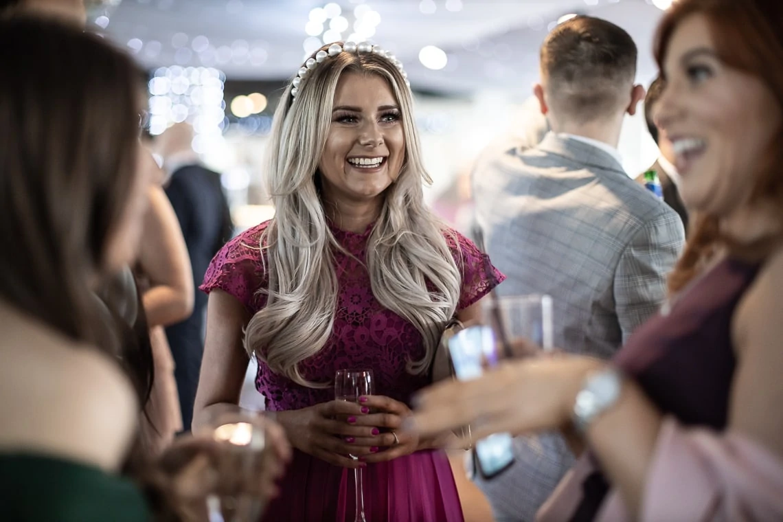 Woman in pink dress with a headband laughs joyfully while holding a drink at a social gathering with others around her.